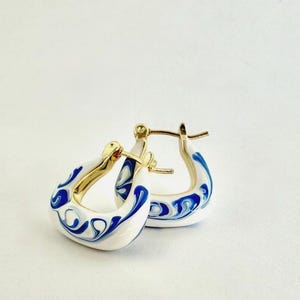 May include: A pair of gold-tone hoop earrings with a white enamel finish. The earrings feature a blue swirled pattern on the outer surface. The inner portion of the hoops is gold. The earrings are displayed on a white background.