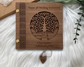 May include: A wooden anniversary gift set. The square wooden plaque reads "Happy 5th Wedding Anniversary" and features a laser-cut tree design. A heart-shaped keychain with initials is included. The set is on a white fur surface.