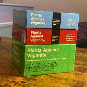 Puede incluir: Tres cajas del juego de cartas "Plants Against Veganity." Las cajas est&aacute;n apiladas una encima de la otra. La caja superior es verde y dice "Plants Against Veganity." La caja del medio es roja y dice "Plants Against Veganity." La caja inferior es azul y dice "Plants Against Veganity." Las cajas est&aacute;n todas decoradas con hojas y flores verdes.