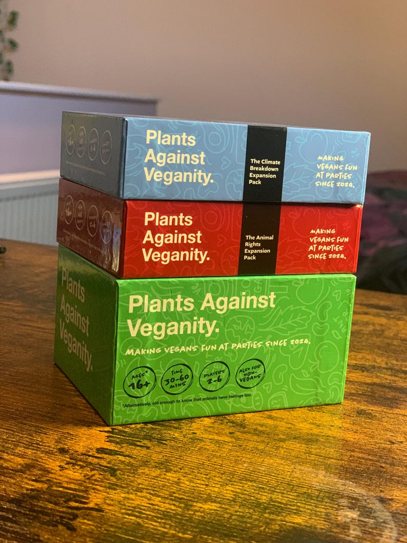 Puede incluir: Tres cajas del juego de cartas "Plants Against Veganity." Las cajas est&aacute;n apiladas una encima de la otra. La caja superior es verde y dice "Plants Against Veganity." La caja del medio es roja y dice "Plants Against Veganity." La caja inferior es azul y dice "Plants Against Veganity." Las cajas est&aacute;n todas decoradas con hojas y flores verdes.