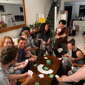 Puede incluir: Un grupo de amigos jugando a un juego de cartas alrededor de una mesa de caf&eacute;. La mesa est&aacute; cubierta de cartas y una peque&ntilde;a caja. Los amigos se r&iacute;en y se divierten.