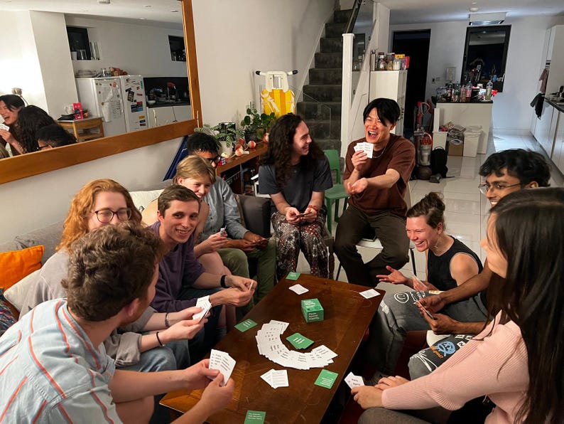 Puede incluir: Un grupo de amigos jugando a un juego de cartas alrededor de una mesa de caf&eacute;. La mesa est&aacute; cubierta de cartas y una peque&ntilde;a caja. Los amigos se r&iacute;en y se divierten.