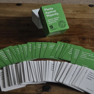 Puede incluir: Una caja verde con el texto "Plants Against Veganity" y el texto "Make vegan friends, lose at parties, once you're done." Un abanico de cartas verdes y blancas con texto en ellas. Las cartas son para un juego.