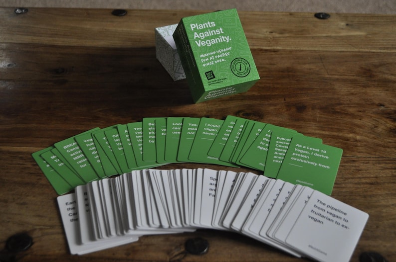 Puede incluir: Una caja verde con el texto "Plants Against Veganity" y el texto "Make vegan friends, lose at parties, once you're done." Un abanico de cartas verdes y blancas con texto en ellas. Las cartas son para un juego.
