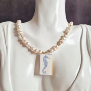 Könnte beinhalten: Eine weiße Perlenkette mit einem blauen und weißen Porzellananhänger mit einem Seepferdchen-Motiv.