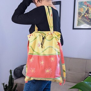 Peut inclure: Une personne portant une chemise noire à manches longues et un jean bleu porte un sac fourre-tout jaune et rose avec une fermeture à cordon et des bretelles réglables.