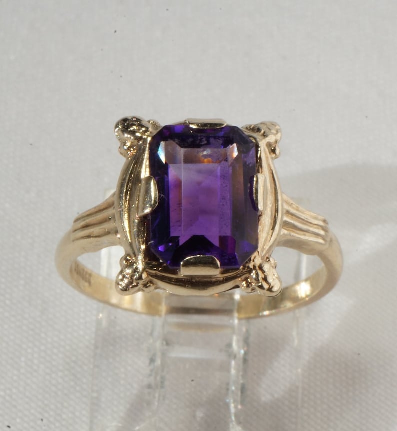 Pu&ograve; includere: Un anello d'oro con una grande pietra preziosa rettangolare viola scuro. L'anello presenta un design ornato attorno alla pietra preziosa e sulle spalle della fascia. L'anello &egrave; esposto su un supporto trasparente.