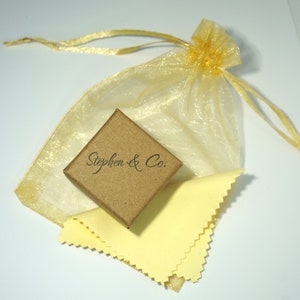 Pu&ograve; includere: Una piccola scatola quadrata di cartone marrone con la scritta "Stephen & Co." in corsivo. La scatola &egrave; accanto a un panno per la pulizia giallo e a un sacchetto regalo in organza dorata con chiusura a coulisse. Lo sfondo &egrave; bianco.