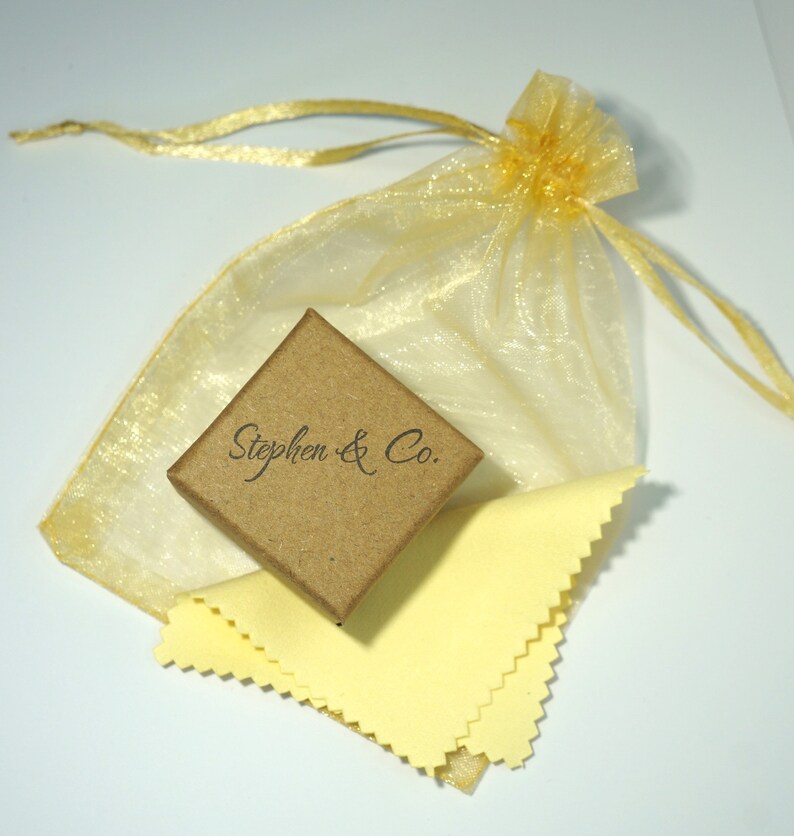 Pu&ograve; includere: Una piccola scatola quadrata di cartone marrone con la scritta "Stephen & Co." in corsivo. La scatola &egrave; accanto a un panno per la pulizia giallo e a un sacchetto regalo in organza dorata con chiusura a coulisse. Lo sfondo &egrave; bianco.