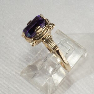 Pu&ograve; includere: Un anello d'oro con una grande gemma rettangolare di colore viola scuro. L'anello ha un design ornato con elementi decorativi sull'incastonatura e sulle spalle. L'anello &egrave; esposto su un supporto di plastica trasparente.