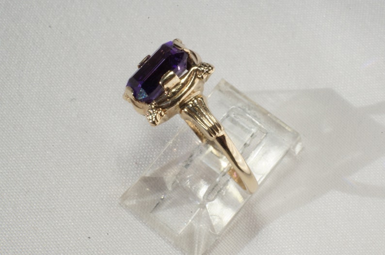 Pu&ograve; includere: Un anello d'oro con una grande gemma rettangolare di colore viola scuro. L'anello ha un design ornato con elementi decorativi sull'incastonatura e sulle spalle. L'anello &egrave; esposto su un supporto di plastica trasparente.