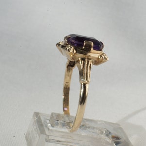 Pu&ograve; includere: Un anello d'oro con una grande pietra preziosa rettangolare viola scuro. L'anello ha un'incastonatura ornata con elementi decorativi attorno alla pietra. La fascia &egrave; in oro lucido. L'anello &egrave; esposto su un supporto trasparente.