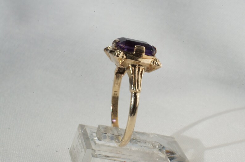 Pu&ograve; includere: Un anello d'oro con una grande pietra preziosa rettangolare viola scuro. L'anello ha un'incastonatura ornata con elementi decorativi attorno alla pietra. La fascia &egrave; in oro lucido. L'anello &egrave; esposto su un supporto trasparente.
