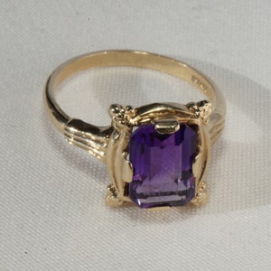 Pu&ograve; includere: Un anello d'oro con una pietra preziosa rettangolare, viola intenso. L'anello ha una montatura ornata con elementi decorativi sulle spalle. La fascia &egrave; in oro lucido semplice. L'anello &egrave; esposto su una superficie bianca.