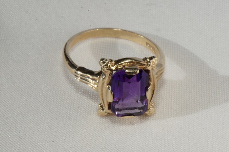 Pu&ograve; includere: Un anello d'oro con una pietra preziosa rettangolare, viola intenso. L'anello ha una montatura ornata con elementi decorativi sulle spalle. La fascia &egrave; in oro lucido semplice. L'anello &egrave; esposto su una superficie bianca.