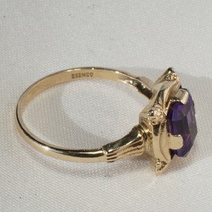 Pu&ograve; includere: Un anello d'oro con una grande pietra preziosa viola taglio ovale. L'anello ha una montatura ornata con dettagli decorativi. La fascia &egrave; sottile e liscia, e il marchio "ESEMCO" &egrave; visibile. L'anello &egrave; posto su uno sfondo bianco.