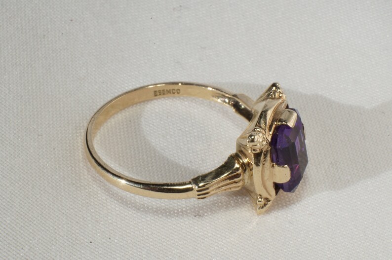 Pu&ograve; includere: Un anello d'oro con una grande pietra preziosa viola taglio ovale. L'anello ha una montatura ornata con dettagli decorativi. La fascia &egrave; sottile e liscia, e il marchio "ESEMCO" &egrave; visibile. L'anello &egrave; posto su uno sfondo bianco.
