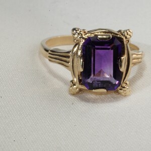 Pu&ograve; includere: Un anello d'oro con una grande ametista rettangolare viola scuro. L'anello ha un'incastonatura ornata con elementi decorativi sulle spalle e intorno alla gemma. L'anello &egrave; posto su uno sfondo bianco.