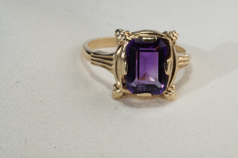 Pu&ograve; includere: Un anello d'oro con una grande ametista rettangolare viola scuro. L'anello ha un'incastonatura ornata con elementi decorativi sulle spalle e intorno alla gemma. L'anello &egrave; posto su uno sfondo bianco.