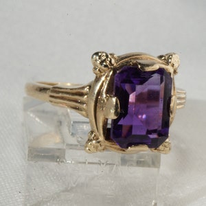 Pu&ograve; includere: Un anello d'oro con una grande pietra preziosa rettangolare viola scuro. L'anello ha un'incastonatura ornata con elementi decorativi attorno alla pietra e sulla fascia. L'anello &egrave; esposto su un supporto trasparente.