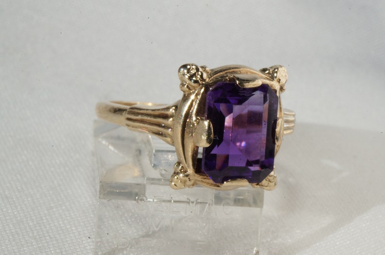 Pu&ograve; includere: Un anello d'oro con una grande pietra preziosa rettangolare viola scuro. L'anello ha un'incastonatura ornata con elementi decorativi attorno alla pietra e sulla fascia. L'anello &egrave; esposto su un supporto trasparente.