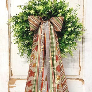 May include: A round wreath made of dense green foliage featuring a large decorative bow at the top. The bow has long, cascading ribbons in various patterns including brown and green stripes, a brown gingham check, and a tan fabric with maple leaves.