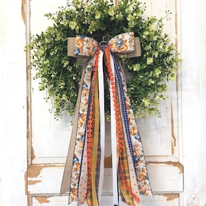 May include: A circular wreath made of green boxwood foliage, adorned with a large decorative bow featuring a pumpkin print. Long, cascading ribbons in various textures and patterns, including plaid, solid orange, white, and blue, hang down.