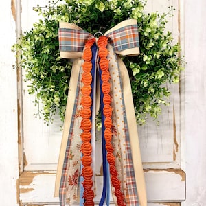 May include: A decorative wreath featuring lush green foliage adorned with a large multi-layered bow. The bow includes plaid ribbons in blue, orange, and cream, alongside long streamers featuring orange pumpkin trim and autumn leaf patterns.