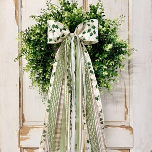 May include: A round, green wreath with a large bow made of fabric with white floral and green leaf patterns. The wreath has long, decorative ribbons in various shades of green and white. The wreath is hanging on a white, distressed door.