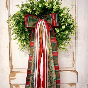 May include: A round wreath made of dense green foliage featuring a large decorative bow at the center. The bow is constructed from multiple ribbons in red, green, and tan, displaying plaid, striped, and botanical patterns that hang downward.