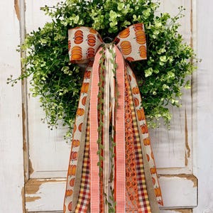 May include: A circular wreath made of green foliage featuring a large decorative bow at the top. The bow and long, trailing ribbons are crafted from various fabrics, including pumpkin-patterned linen, orange gingham, plaid, and lace textures.