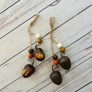 May include: Two acorn ornaments with brown, orange, and white beads strung on twine. The acorns have a metallic finish and are designed for hanging. The background is a light wood surface.