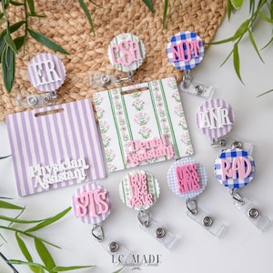 May include: A collection of medical-themed badge reels and name tags. The reels feature various designs, including gingham, stripes, and floral patterns, with phrases like "ER," "RAD," and "Physician Assistant." The color palette includes pink, blue, and white.