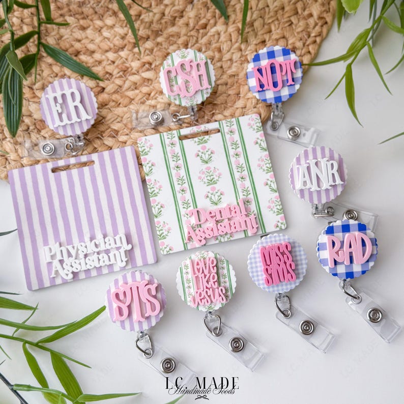 May include: A collection of medical-themed badge reels and name tags. The reels feature various designs, including gingham, stripes, and floral patterns, with phrases like "ER," "RAD," and "Physician Assistant." The color palette includes pink, blue, and white.