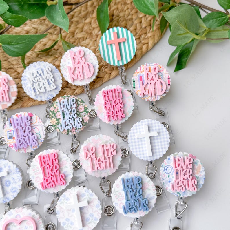May include: A collection of decorative badge reels with various designs and inspirational phrases. The reels feature phrases like "Be Kind," "Love Like Jesus," and "Faith Over Fear." The designs include crosses, floral patterns, and striped backgrounds, in shades of pink, blue, and white.