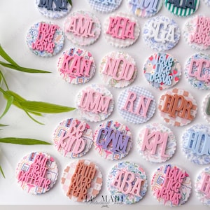 May include: A collection of decorative round embellishments with various phrases and initials in pink, purple, and brown. The embellishments have a scalloped edge and patterned backgrounds, including floral, gingham, and plaid designs. The text "LC MADE Handmade Goods" is visible.