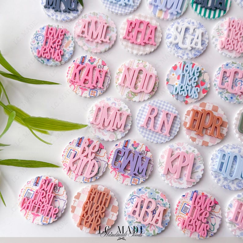 May include: A collection of decorative round embellishments with various phrases and initials in pink, purple, and brown. The embellishments have a scalloped edge and patterned backgrounds, including floral, gingham, and plaid designs. The text "LC MADE Handmade Goods" is visible.
