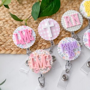 May include: A collection of decorative badge reels with various designs and text. The reels feature floral patterns, gingham, and text such as "LME", "IMG", "RN", and "PSALM 23". Each reel has a clear plastic strap and a metal clip.