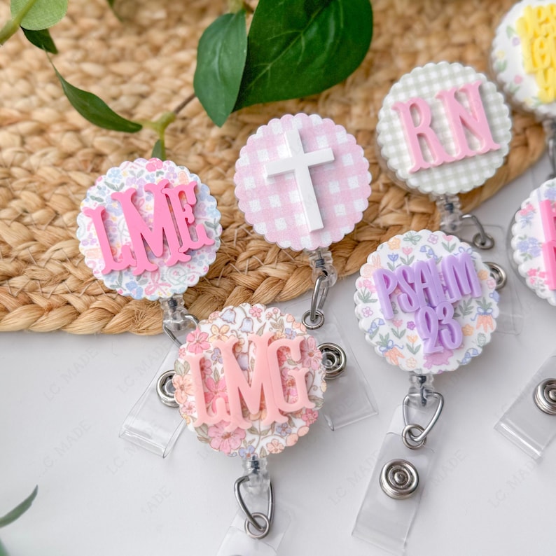 May include: A collection of decorative badge reels with various designs and text. The reels feature floral patterns, gingham, and text such as "LME", "IMG", "RN", and "PSALM 23". Each reel has a clear plastic strap and a metal clip.