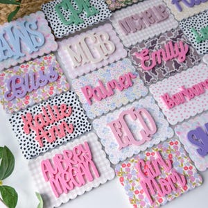 May include: A collection of rectangular scalloped-edge plaques featuring various patterns including floral, gingham, and polka dots. Each plaque displays raised, three-dimensional lettering in diverse colors, spelling out names and monograms.