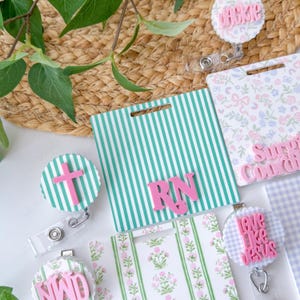 May include: A collection of badge reels and name tag holders with various designs. The badge reels feature pink crosses and text, while the name tag holders have striped and floral patterns. The letters "RN" are displayed on a green and white striped background.