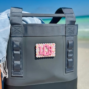 May include: A dark gray cooler bag with a handle and straps, featuring a pink monogrammed patch with the letters "LCB". The patch has a floral pattern. A blue and white striped towel is visible inside the cooler.