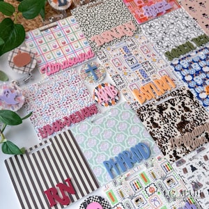 May include: A collection of square badge reels featuring various patterns including plaid, leopard print, cow print, stripes, and floral designs. Several reels display raised lettering such as Ultrasound, BSNRN, Labor+Delivery, BSTRN, PHARM, and Respiratory Care.