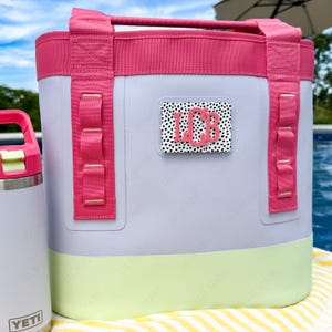 May include: A light purple cooler bag with a bright pink top trim and handles, a light green base, and a rectangular patch featuring a black and white dalmatian-style dot pattern with the pink monogram letters LCB centered on the front.