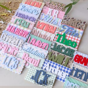May include: A collection of personalized name tags in various colors and fonts. Each tag features a name in a 3D style, set against patterned backgrounds like floral, plaid, and gingham. The tags are designed for keychains.
