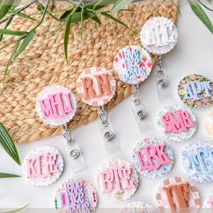 May include: A collection of decorative badge reels with various designs and initials. The reels feature floral, plaid, and patterned backgrounds with raised lettering in pink, peach, and white. Some reels have the text "love like Jesus".