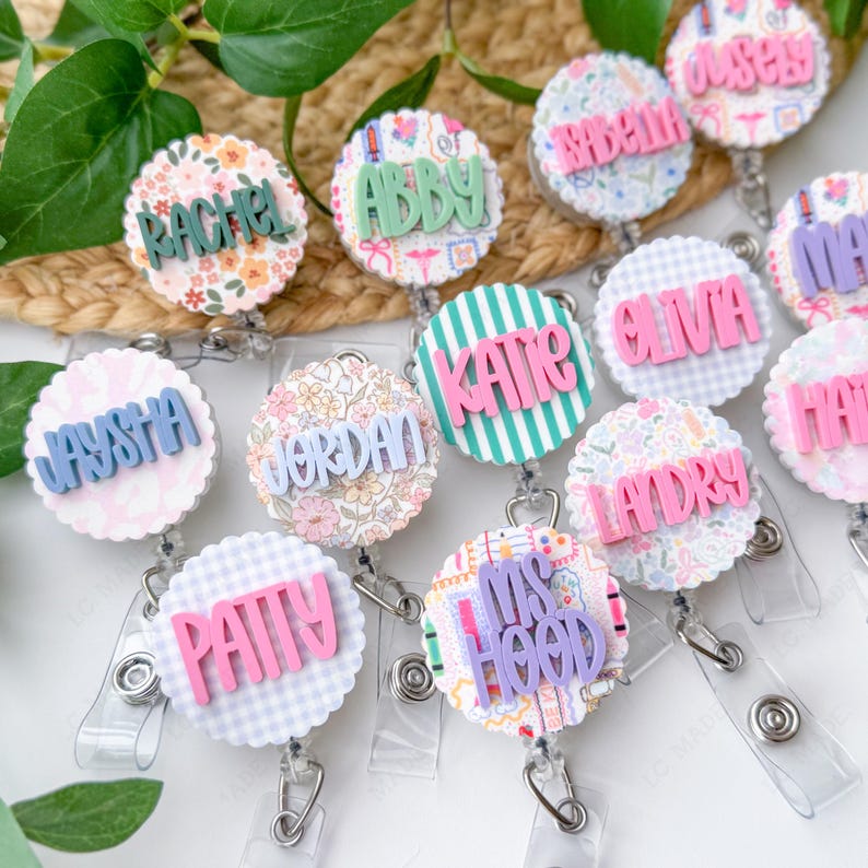 May include: A collection of personalized badge reels with names like Rachel, Abby, and Katie. Each reel features a unique patterned background, including floral, striped, and gingham designs, with the name in a contrasting color. The reels have a scalloped edge.