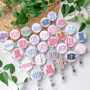 May include: A collection of personalized badge reels with various initials and phrases. The round, scalloped-edge designs feature pastel colors, floral patterns, and striped backgrounds. Each reel has a clear plastic strap and metal clip.