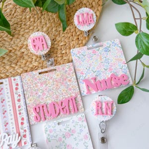 May include: A collection of floral-patterned badge holders and matching badge reels. The badge holders feature the words "Nurse" and "Student" in pink, along with initials on the reels. The background is a woven texture with green leaves.