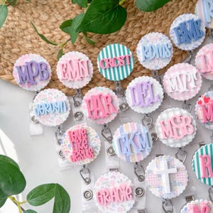 May include: A collection of personalized badge reels with various designs and text. The reels feature floral, striped, and patterned backgrounds with raised lettering in pastel colors. Some reels display initials, while others have phrases like "Love Like Jesus" and "Grace".